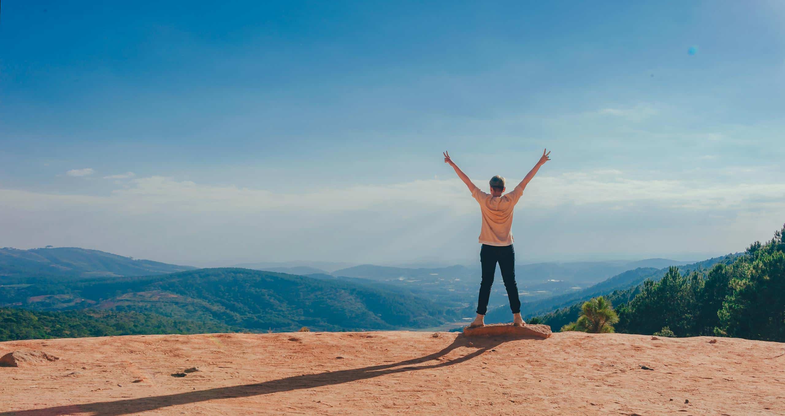 A man raises his arms in triumph on a rocky mountain summit overlooking a vast landscape