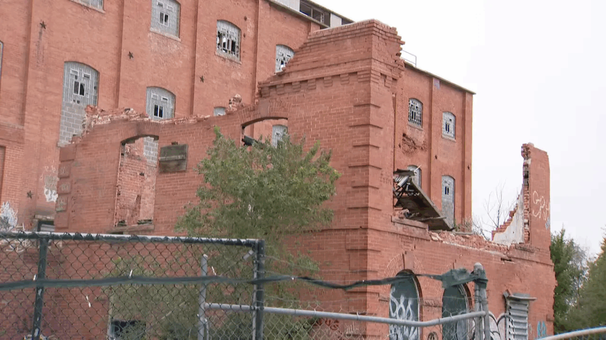 Wide view of the crumbling brick sugar factory building behind a chain-link fence with damaged sections and boarded openings.